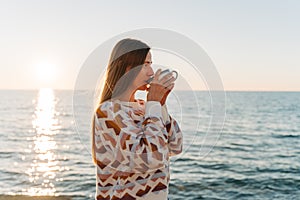 Young beautiful girl in cozy sweater drinking coffee while enjoying winter sun on seaside shore during mild sunset. Cute
