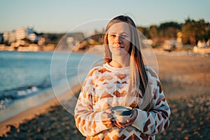 Young beautiful girl in cozy sweater holding coffee cup while enjoying winter sun on seaside shore during mild sunset