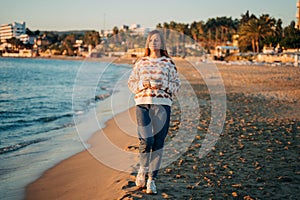 Young beautiful girl in cozy sweater holding coffee cup while enjoying winter sun on seaside shore during mild sunset