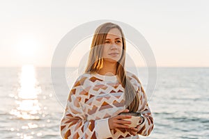 Young beautiful girl in cozy sweater holding coffee cup while enjoying winter sun on seaside shore during mild sunset