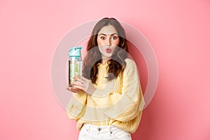 Young beautiful girl with curly hairstyle, pucker lips and looks excited, showing lemon water bottle drink, standing