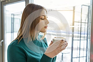 Young beautiful girl drinking coffee, standing in profile, looking out the window. Autumn winter season in coffee shop