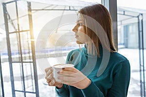 Young beautiful girl drinking coffee, standing in profile, looking out the window. Autumn winter season in coffee shop