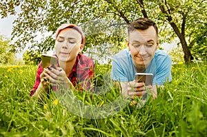 Young beautiful girl and guy lies on green grass and dandelions and talking smartphones