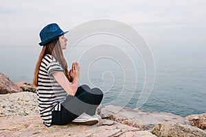 Young beautiful girl in a hat practicing yoga and meditating on stones next to the sea. Recreation. Vacation. Relaxation