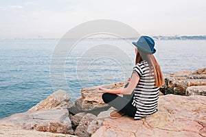 Young beautiful girl in a hat practicing yoga and meditating on stones next to the sea. Recreation. Vacation. Relaxation