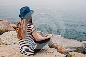 Young beautiful girl in a hat practicing yoga and meditating on stones next to the sea. Recreation. Vacation. Relaxation