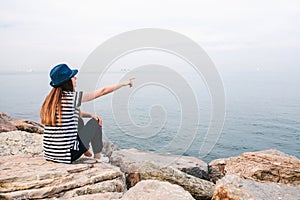 Young beautiful girl in a hat sits on the rocks at the seaside and shows her hand to the distance. Rest, vacation