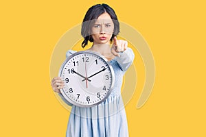 Young beautiful girl holding big clock pointing with finger to the camera and to you, confident gesture looking serious