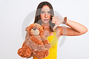 Young beautiful girl holding cute teddy bear standing over isolated white background with angry face, negative sign showing