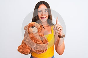 Young beautiful girl holding cute teddy bear standing over isolated white background surprised with an idea or question pointing