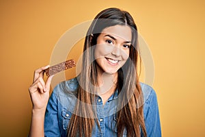 Young beautiful girl holding healthy protein bar standing over isolated yellow background with a happy face standing and smiling