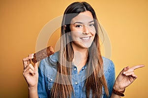 Young beautiful girl holding healthy protein bar standing over isolated yellow background very happy pointing with hand and finger