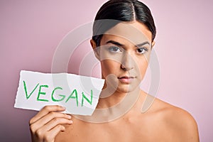 Young beautiful girl holding paper with vegan message over isolated pink background with a confident expression on smart face