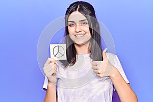 Young beautiful girl holding peace symbol reminder smiling happy and positive, thumb up doing excellent and approval sign