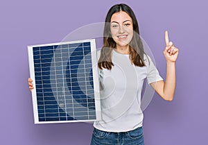 Young beautiful girl holding photovoltaic solar panel surprised with an idea or question pointing finger with happy face, number