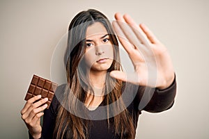 Young beautiful girl holding sweet bar of chocolate over isolated white background with open hand doing stop sign with serious and