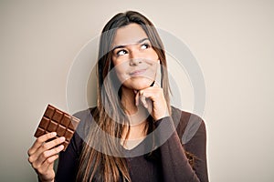 Young beautiful girl holding sweet bar of chocolate over isolated white background serious face thinking about question, very