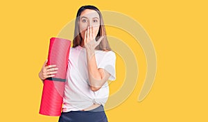 Young beautiful girl holding yoga mat covering mouth with hand, shocked and afraid for mistake