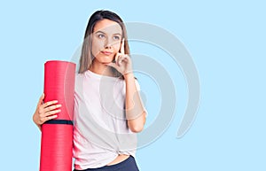 Young beautiful girl holding yoga mat serious face thinking about question with hand on chin, thoughtful about confusing idea