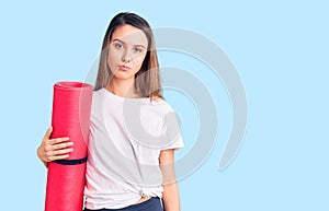 Young beautiful girl holding yoga mat thinking attitude and sober expression looking self confident