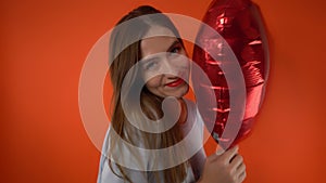 young beautiful girl holds heart-shaped balloon on red background in her hand and sends air kiss to camera. Valentine's