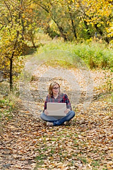 Young beautiful girl holds a laptop in her hands, sitting on fallen leaves in the forest with her legs crossed, pondering