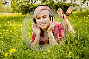 Young beautiful girl listening to music on headphones and lies on a green grass and dandelions