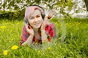 Young beautiful girl listening to music on headphones and lies on a green grass and dandelions