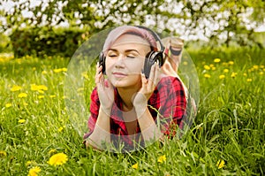 Young beautiful girl listening to music on headphones and lies on a green grass and dandelions