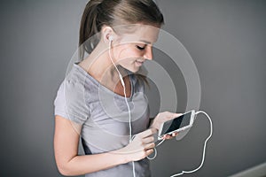 Young beautiful girl listening to music with headphones while showing blank screen mobile phone over gray.