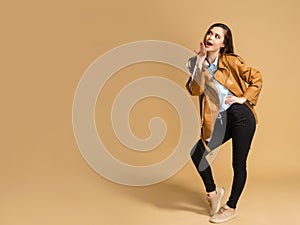 Young beautiful girl with long curly hair in a brown leather jacket covers her mouth with her hand on an orange studio background