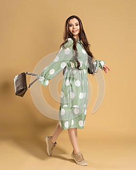 Young beautiful girl with long curly hair in a dress with polka dots with a bag in her hands posing on an orange studio background