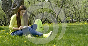 Young beautiful girl with long dark hair sitting in nature on a green field with dandelions under a tree, reading a book