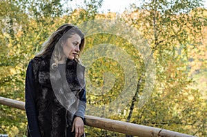 Young beautiful girl with long hair, in a fur jacket, stands on a wooden railing on a bright sunny day while walking in the autumn