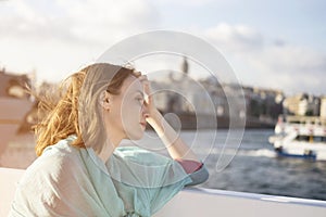 Young beautiful girl in pink and blue dress sitting on boat or ferry at sunset and thoughtfully looks at the sea.