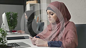Young beautiful girl in pink hijab sits in office and uses smartphone. Girl in black hijab in the background. Arab women