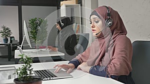 A young beautiful girl in a pink hijab is talking on the headset, answering calls in call center. Arab women in the