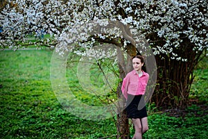 Young beautiful girl in a pink shirt standing under blossoming apple tree and enjoying a sunny day.