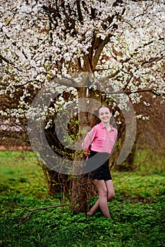 Young beautiful girl in a pink shirt standing under blossoming apple tree and enjoying a sunny day.