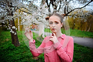 Young beautiful girl in a pink shirt standing under blossoming apple tree and enjoying a sunny day.