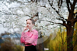 Young beautiful girl in a pink shirt standing under blossoming apple tree and enjoying a sunny day.