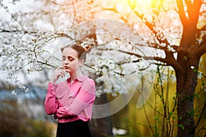 Young beautiful girl in a pink shirt standing under blossoming apple tree and enjoying a sunny day.