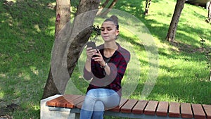 Young beautiful girl in a plaid shirt sits on a bench in the park, actively rewritten in internet chat.