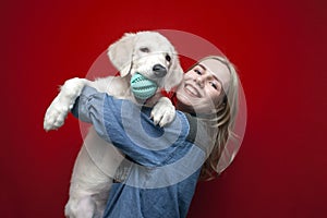 Young beautiful girl plays with a golden retriever puppy on a red background, a dog grizzles a toy ball, the hostess hugs a pet