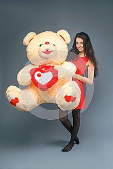 Young beautiful girl in red dress with big teddy bear soft toy happy smiling and playing on grey background