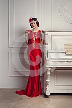 Young beautiful girl in red dress style of the 20`s or 30`s with glass of martini near the piano. Vintage style beautiful woman.