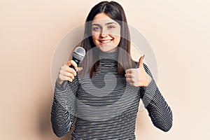 Young beautiful girl singing song using microphone smiling happy and positive, thumb up doing excellent and approval sign