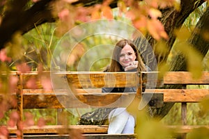 A young beautiful girl sits on a bench in an autumn park and looks into the frame, smiling.