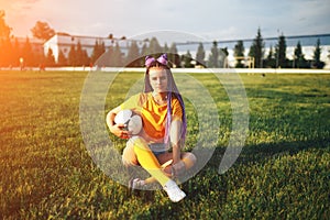 Young beautiful girl sitting on a lawn holding a soccer ball in her hand in a yellow T-shirt and open socks